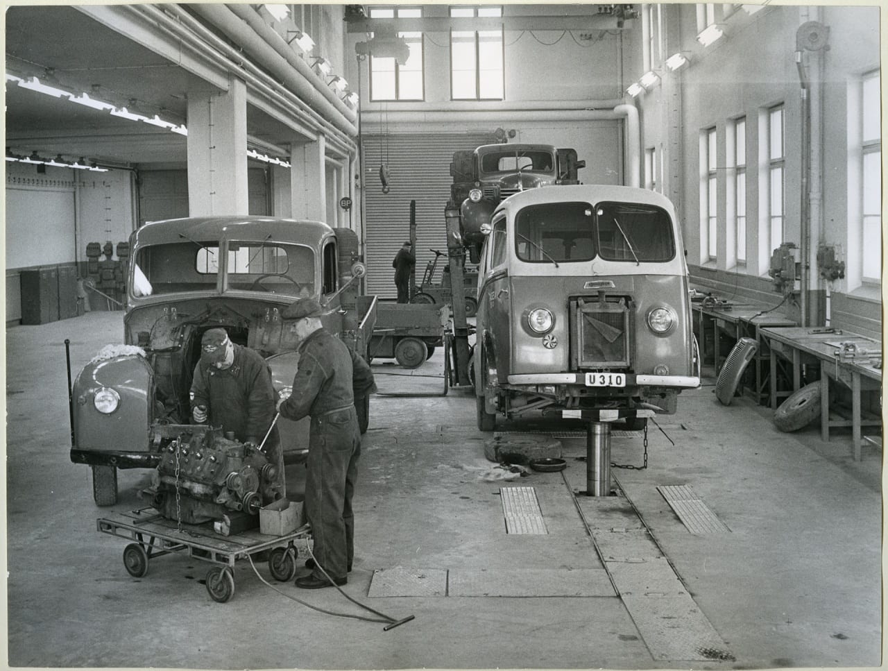 Aseas bilverkstad i Viksängshuset med hydraliska billyftar år 1956. Tre bilar står inne i verkstaden och tre verkstadsarbetare arbetar med dem.